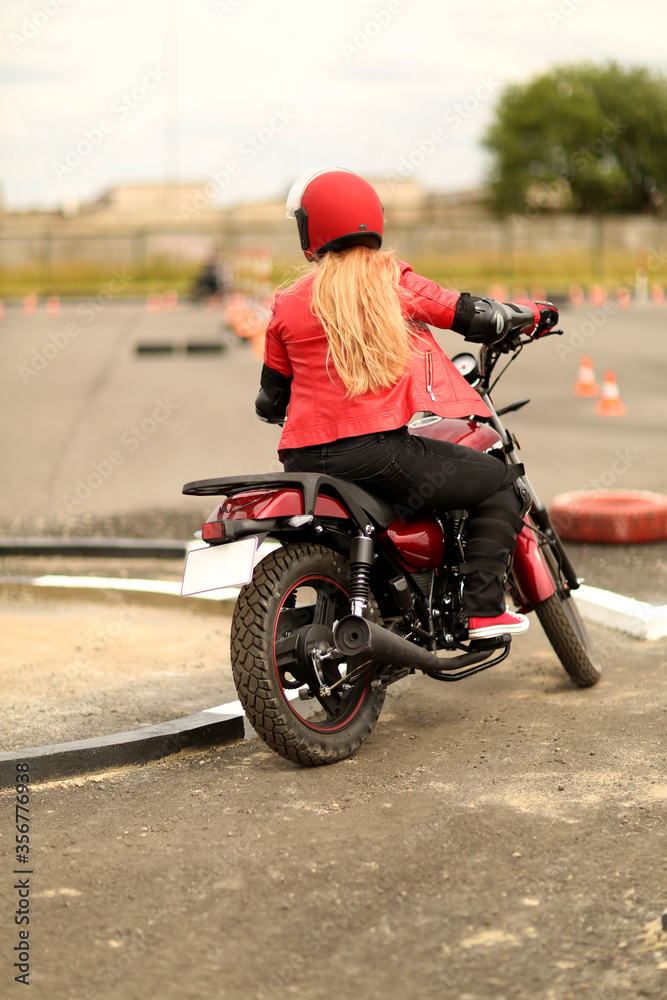 A girl motorcyclist in a red jacket passes an exam in a driving school. A woman rides a bike. Woman in a red helmet rides a motorcycle.