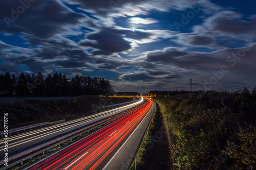 Aerial view of highway at night. Rear red and front white light trail visible on road. Long exposure photography. Full moon and illuminated clouds on the sky