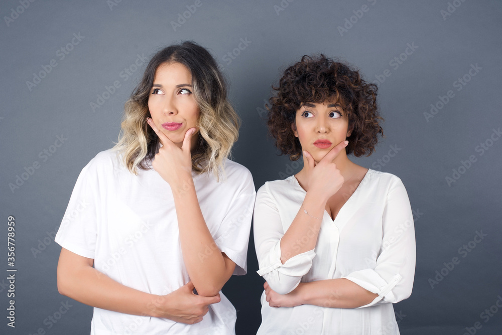 Isolated portrait of stylish young European woman with hand under chin and looking sideways with doubtful and skeptical expression, worry and doubt. Standing indoors over gray background. Isolated portrait of stylish young European woman with hand under chin and looking sideways with doubtful and skeptical expression, worry and doubt. Standing indoors over gray background.