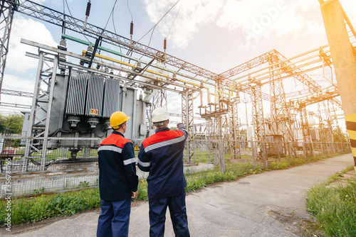 Two specialist electrical substation engineers inspect modern high-voltage equipment during sunset. Energy. Industry