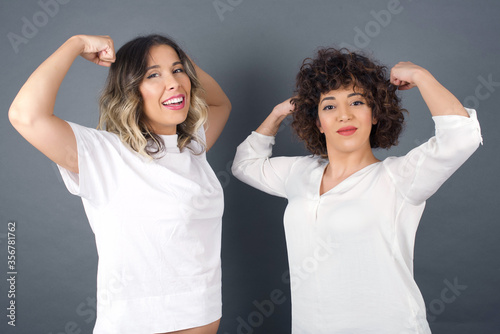 Waist up shot of caucasian woman raises arms to show her muscles feels confident in victory, looks strong and independent, smiles positively at camera, stands against gray background. Sport concept.