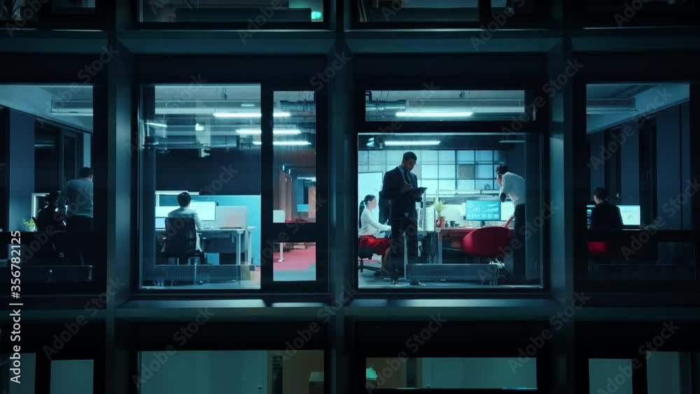 Footage from Outside the Window: Businessmen and Businesswomen Working in the Office. Managers and Specialists Doing Financial Business in the Evening. Employees Work on Computers and Delegate Tasks.