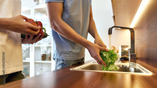 Man s hands washing lettuce in modern kitchen sink before cooking, preparing a meal. Woman helping him, holding other vegetables. Vegetarianism, healthy food, hygiene concept