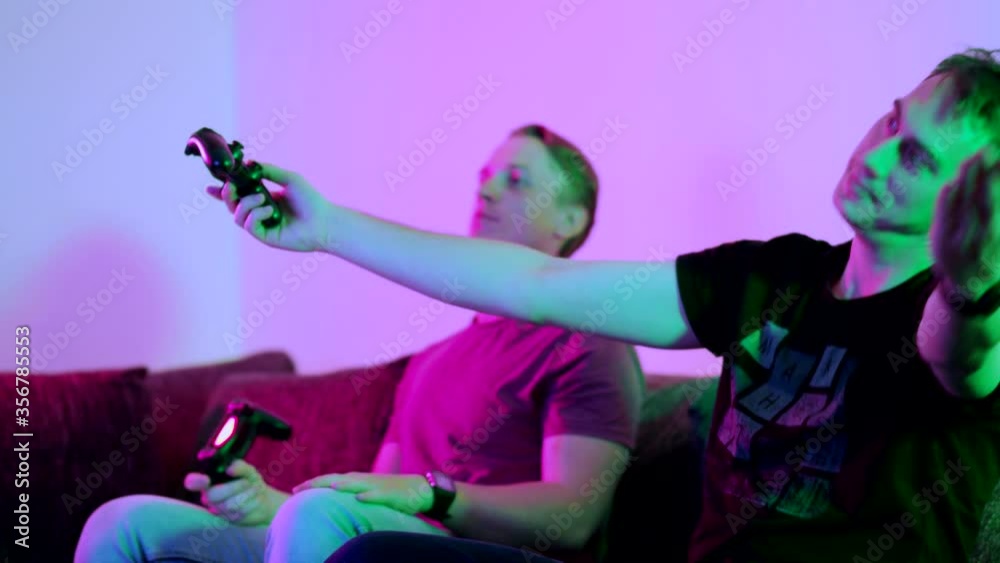 Two happy friends playing action video game in the living room sitting on the sofa in 4K VIDEO. Intense competition between two young male players is full of emotions. Two happy friends playing action video game in the living room sitting on the sofa in 4K VIDEO. Intense competition between two young male players is full of emotions.