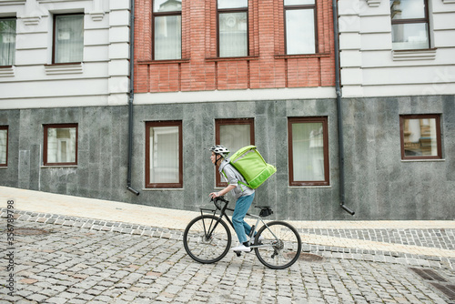 Young delivery woman in helmet with thermo bag or backpack riding a bike along the city, delivering food. Courier, delivery service concept © Svitlana