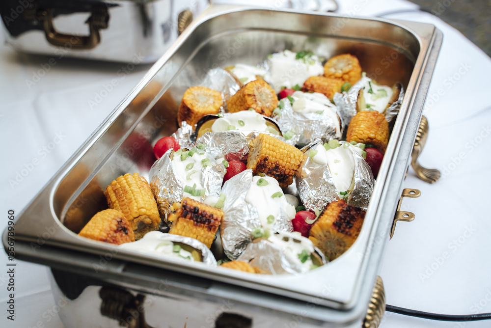 Grilled corn and baked potatoes stuffed with cheese in hotel pan on food warmer. Self-service buffet table. Celebration, party, birthday or wedding Grilled corn and baked potatoes stuffed with cheese in hotel pan on food warmer. Self-service buffet table. Celebration, party, birthday or wedding