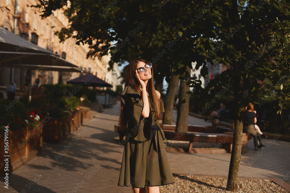 Portrait of a cheerful young woman in dress touching her sunglasses on urban background. Fashionable girl with beautiful brown hair smiling to camera. Portrait of a cheerful young woman in dress touching her sunglasses on urban background. Fashionable girl with beautiful brown hair smiling to camera.
