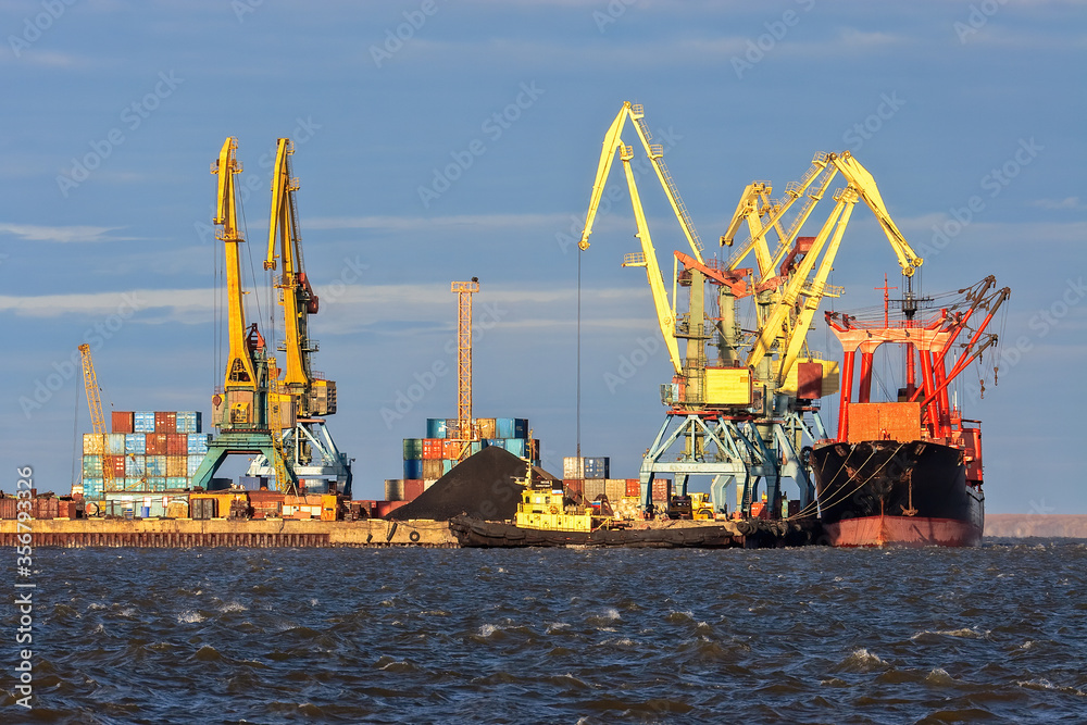 Seaport in the Arctic. A cargo ship near the pier of the sea port. Large port cranes. Logistics, shipping and freight in the Arctic. Stevedore services. Anadyr seaport, Chukotka, Far East Russia.