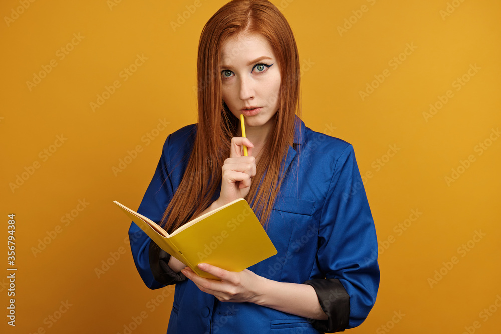 Red-haired girl in a blue jacket stands on a yellow background with a yellow notebook, thoughtfully holding a pencil to her lips Red-haired girl in a blue jacket stands on a yellow background with a yellow notebook, thoughtfully holding a pencil to her lips
