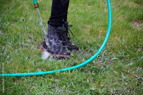 Person rinsing off muddy hiking boots with green hose while standing in grass
