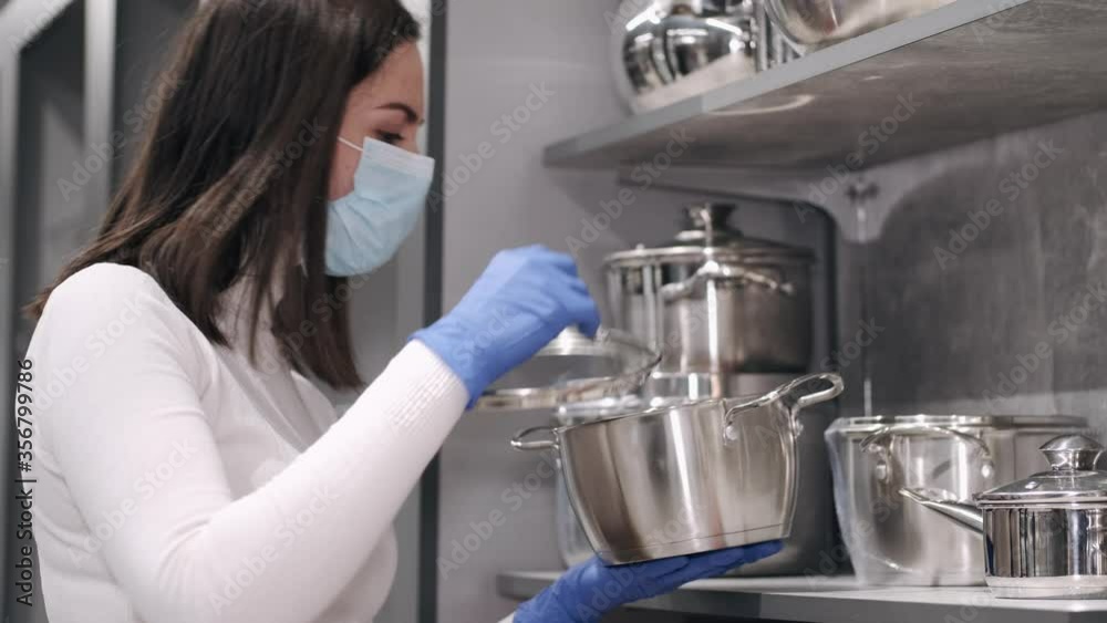 A woman is checking the cleanliness of the tableware. She is inspecting the pans on the shelves. The woman is wearing rubber gloves and a mask. A woman is checking the cleanliness of the tableware. She is inspecting the pans on the shelves. The woman is wearing rubber gloves and a mask.