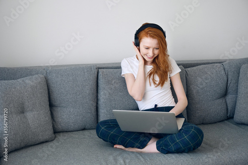 Redhead girl sitting on a sofa in home clothes with a laptop on