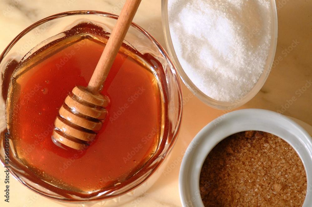 Close up in flat lay angle of a jar of honey next to white sugar and brown sugar bowls.