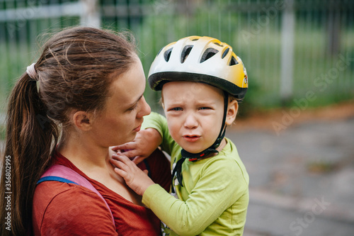 Mom holds a crying todler in her arms. The boy has a helmet on his head. They are on the street. 