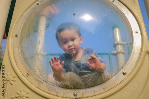 cute little Asian boy sitting on playground touching and looking through clear plastic window 