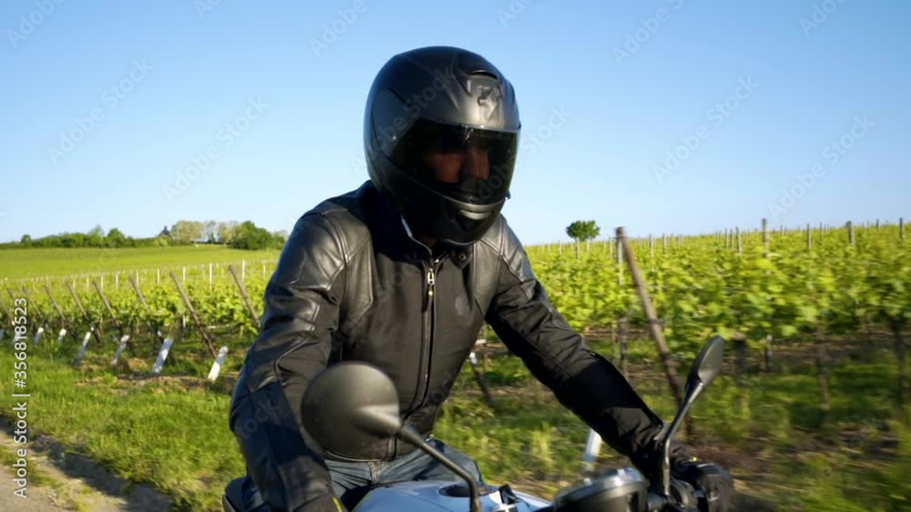 A motorcyclist in full black is driving on an asphalt road between two vineyards on a sunny day with a blue sky - sideview close up tracking shot A motorcyclist in full black is driving on an asphalt road between two vineyards on a sunny day with a blue sky - sideview close up tracking shot