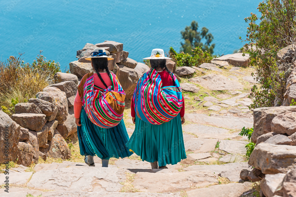 Fototapeta Two indigenous Peruvian Quechua women in traditional clothing walking down the steps to the harbor of Taquile Island, Titicaca Lake, Peru.