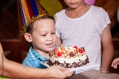 Cute asian child boy is surprising with delicious birthday cake which his mother give to him. Child is blowing candles on birthday cake in the party with fun and happiness.