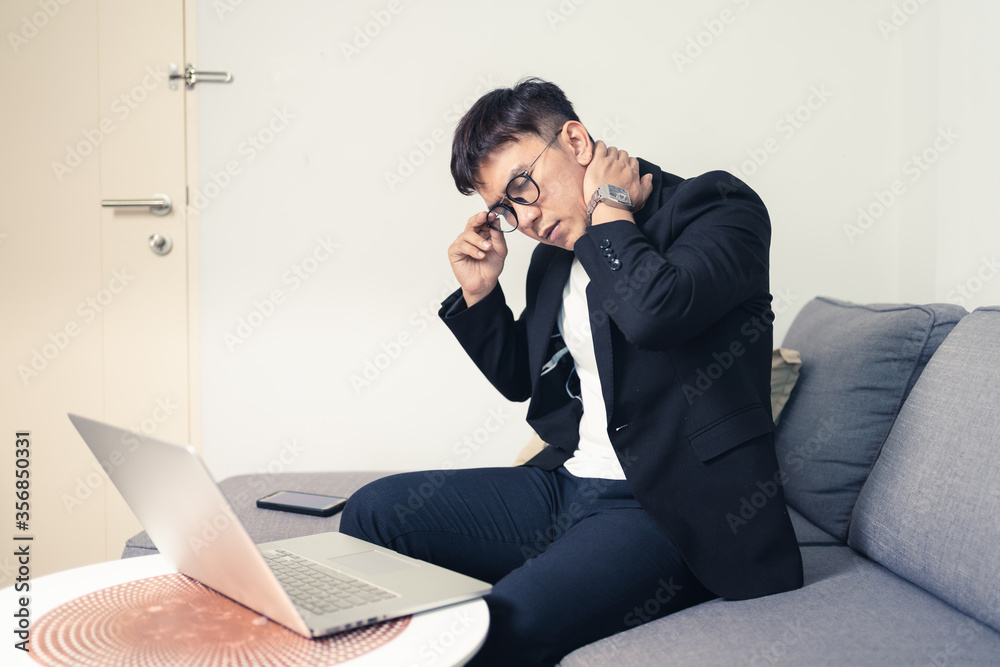 Young Asian business man sitting on sofa and working from home. Man feeling stress and suffering from office syndrome. Tired young entrepreneur manager. Work from home concept Young Asian business man sitting on sofa and working from home. Man feeling stress and suffering from office syndrome. Tired young entrepreneur manager. Work from home concept