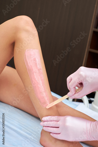 The beautician is preparing for depilation and applying the cream with wax stick on the female legs. Cosmetology procedures at home