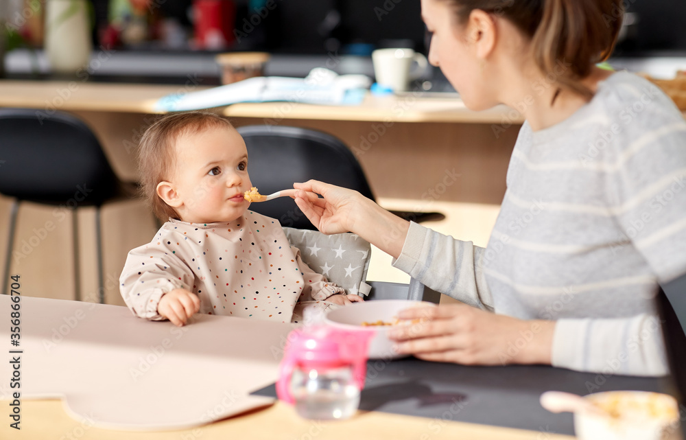 family, eating and people concept - happy mother with puree and spoon feeding little baby girl sitting in highchair at home family, eating and people concept - happy mother with puree and spoon feeding little baby girl sitting in highchair at home