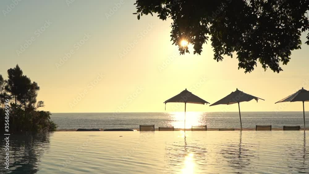 Sunset at the infinity pool area. Beach Umbrellas and deck chairs silhouette in the foreground. Sunlight reflections in pool water Sunset at the infinity pool area. Beach Umbrellas and deck chairs silhouette in the foreground. Sunlight reflections in pool water