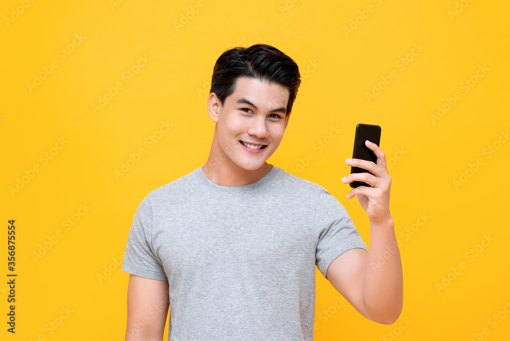 Young smiling handsome Asian man taking a selfie with cellphone isolated yellow studio background Young smiling handsome Asian man taking a selfie with cellphone isolated yellow studio background