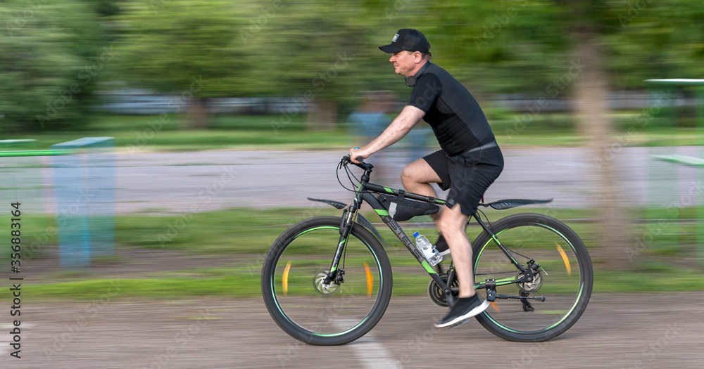 Sterlitamak, Republic of Bashkortostan, Russia - June 10, 2020. Training at the school stadium. A cyclist rushes at breakneck speed against a background of green trees. Sterlitamak, Republic of Bashkortostan, Russia - June 10, 2020. Training at the school stadium. A cyclist rushes at breakneck speed against a background of green trees.