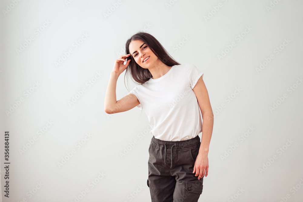 embarrassed brunette with a beautiful smile in a white t-shirt hand straightens her hair on a light background with empty place for text embarrassed brunette with a beautiful smile in a white t-shirt hand straightens her hair on a light background with empty place for text