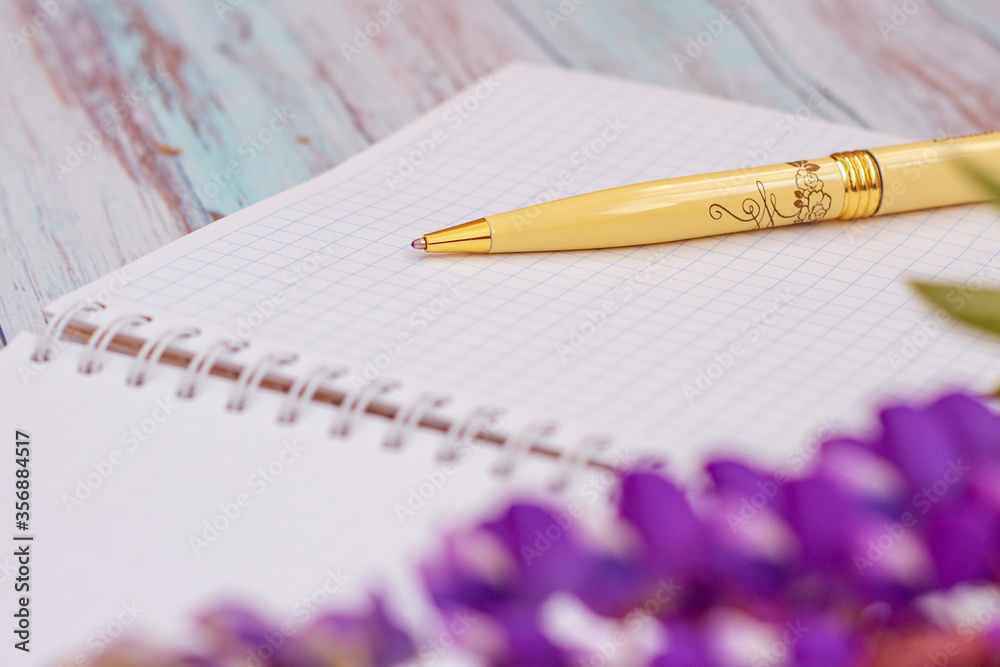 Notebook in a cage with a yellow pen, next to one flower with purple petals and green leaves close-up Notebook in a cage with a yellow pen, next to one flower with purple petals and green leaves close-up