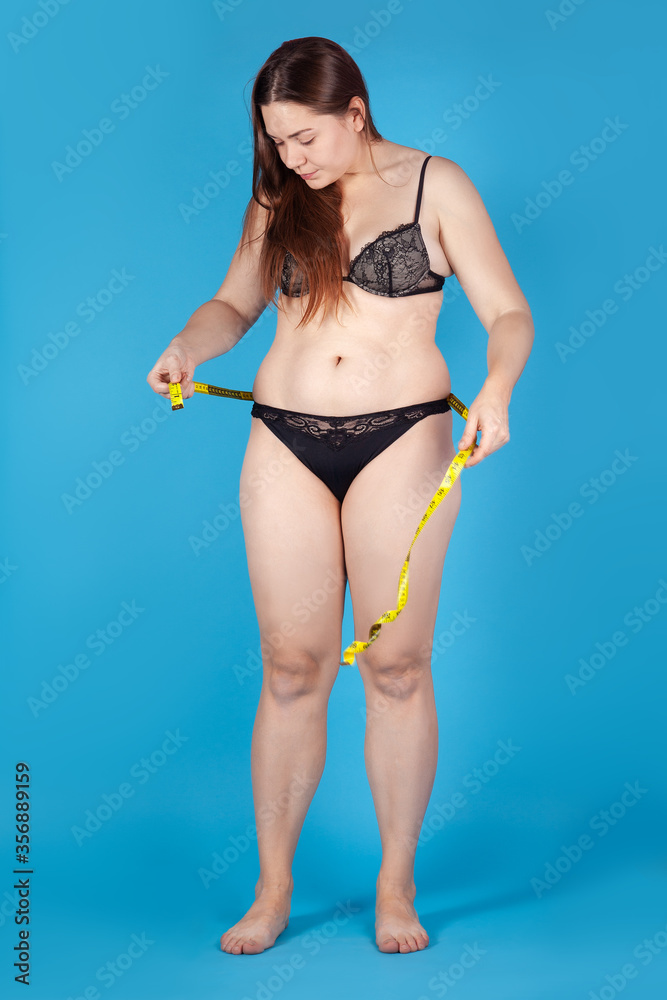 Naklejka premium Young overweighted woman in black underwear measuring her waist and hips with yellow tape. Healthy lifestyle, odd weight or body positive concept. Standing on bright blue background, studio.