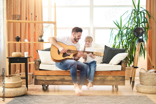 Handsome young man and little cute girl play the guitar at home together. Happy Father's Day! Father and daughter in white t-shirts and jeans sit on couch in the living room. I love you, dad! Family