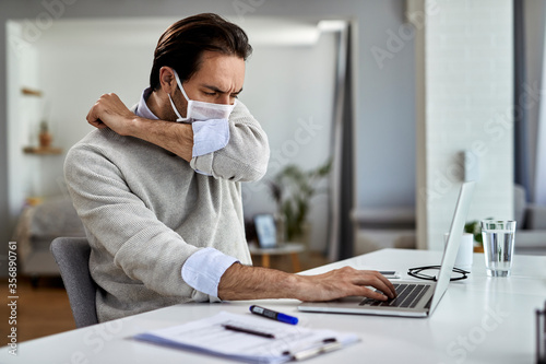 Freelance worker with face mask sneezing into elbow while working on laptop at home.