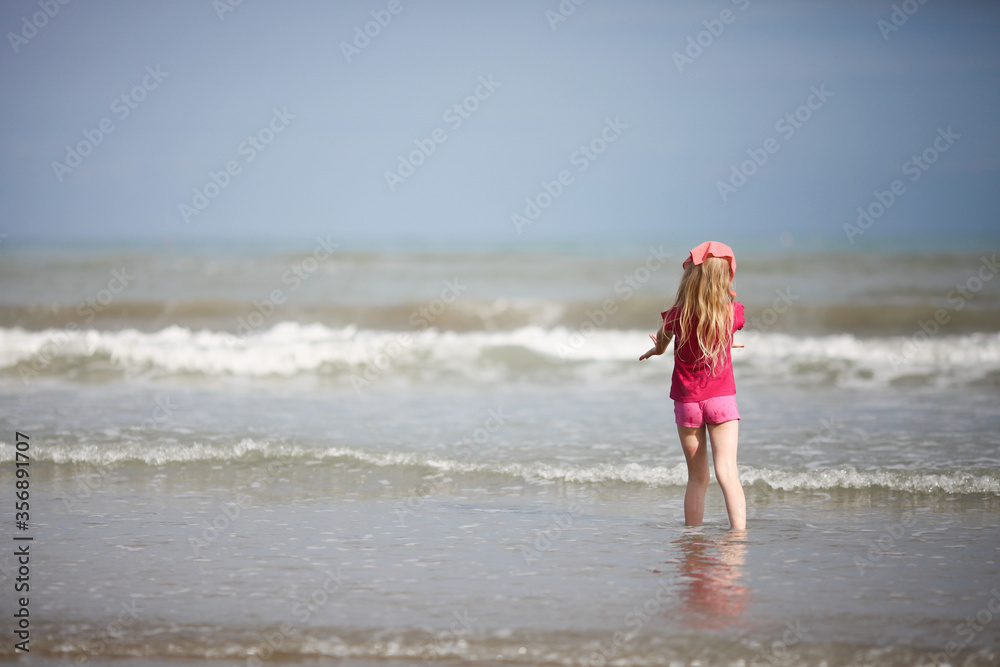 Little girl in kerchief staying facing the sea, hands to the sea. Back view. Lido di Jesolo, Italy. Summer vacation with children concept. Little girl in kerchief staying facing the sea, hands to the sea. Back view. Lido di Jesolo, Italy. Summer vacation with children concept.