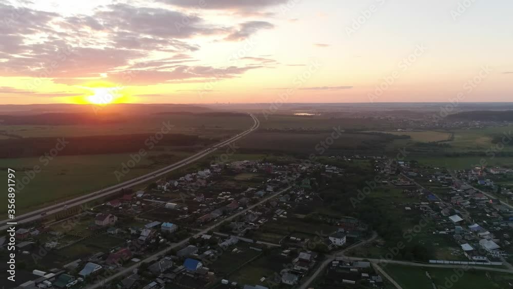 02. Aerial view of evening highway at sunset near the village and field. Cars drive along the highway. The sun falls over the horizon and beautifully illuminates the clouds.. Summer, evening