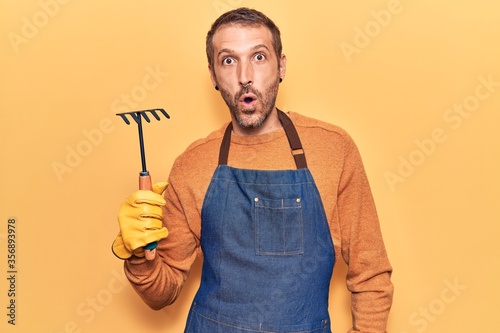 Young handsome man wearing gardener apron and gloves holding rake scared and amazed with open mouth for surprise, disbelief face
