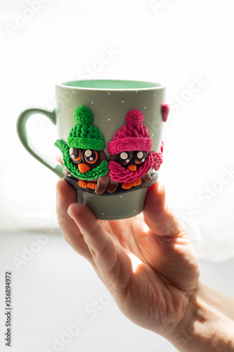 The original mug with decorative figurines made with their own hands. A lovely birthday present. Figures on the cup are fashioned hand made from colored clay. A hand holds cups. High quality photo