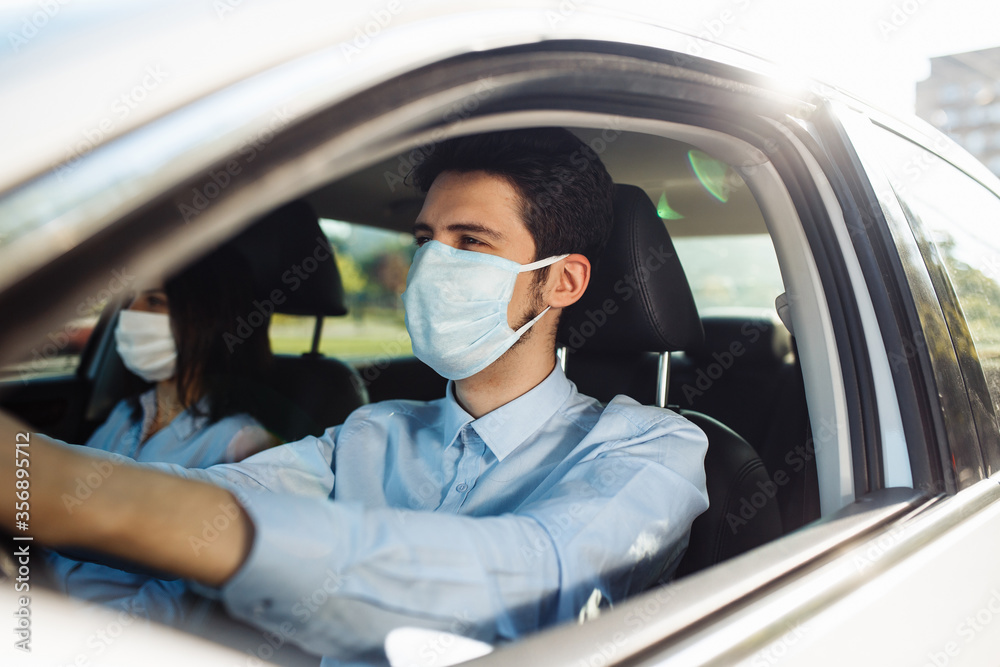 Young man taxi driver wears sterile medical mask in the car. A boy behind the car steering wheel safely driving for business matter keeping social distance. Coronavirus pandemic concept. Young man taxi driver wears sterile medical mask in the car. A boy behind the car steering wheel safely driving for business matter keeping social distance. Coronavirus pandemic concept.
