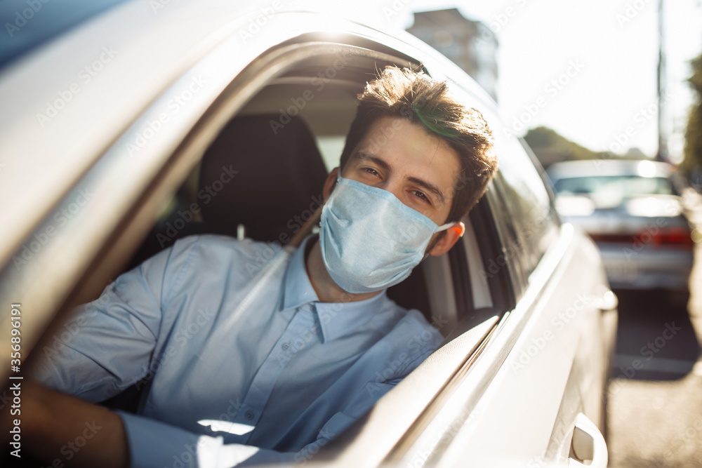 Young man taxi driver looks out of vehicle's window wears sterile medical mask in the car. A boy behind the car steering wheel safely driving keeping social distance. Coronavirus pandemic concept.