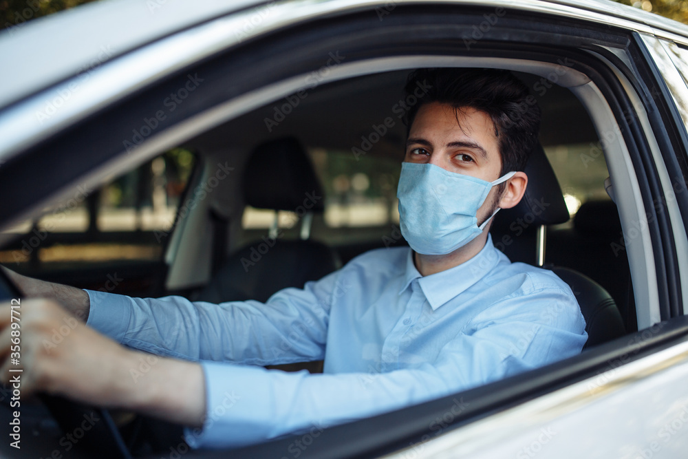 Young man sits behind the steering wheel in the car wearing sterile medical mask. Boy taxi driver works hard during coronavirus outbreak. Social distance, virus spread prevention and treat concept. Young man sits behind the steering wheel in the car wearing sterile medical mask. Boy taxi driver works hard during coronavirus outbreak. Social distance, virus spread prevention and treat concept.