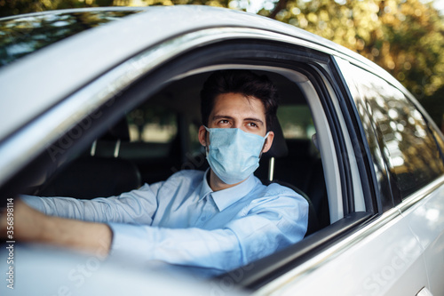 Young man sits behind the steering wheel in the car wearing sterile medical mask. Boy taxi driver works hard during coronavirus outbreak. Social distance, virus spread prevention and treat concept.