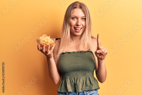 Young blonde woman holding potato chip smiling with an idea or question pointing finger with happy face  number one