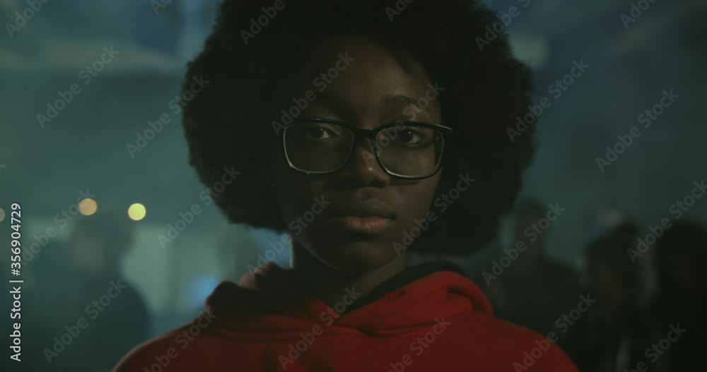 Close up view of young african american girl in glasses looking to camera while standing at demonstration at night street. Crowd of protesters at background. Concept of strike