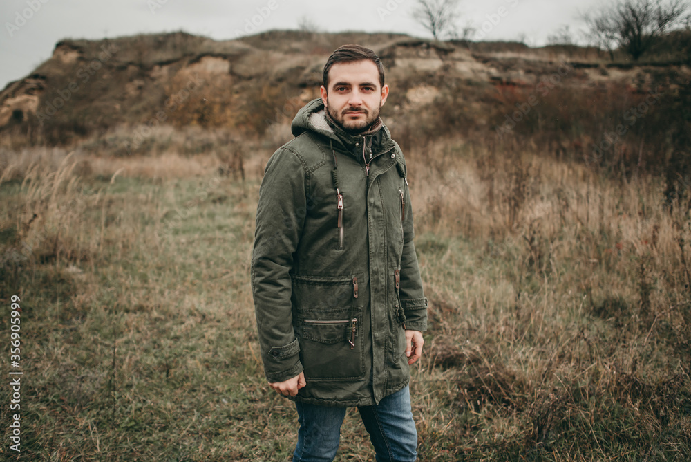 Handsome young stylish man in a green jacket, Scandinavian Christmas sweater and jeans walks in nature. Cloudy weather, spring, dry grass.