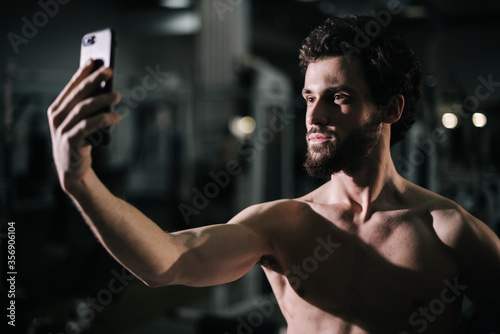 Middle shot portrait of confident bearded young man with muscular wiry naked torso taking selfie photo on smartphone close-up looking camera during sport workout training in modern dark gym. © dikushin