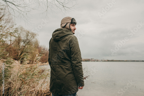 Young man in winter hat, green jacket in nature near the lake in autumn. Travel, cold weather, rain, a guy in a Scandinavian sweater.