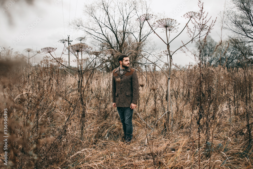 Handsome sadly young stylish man in a jacket and jeans walks on the nature of jubilee dried autumn flowers. Cloudy weather, spring, dry grass Handsome sadly young stylish man in a jacket and jeans walks on the nature of jubilee dried autumn flowers. Cloudy weather, spring, dry grass