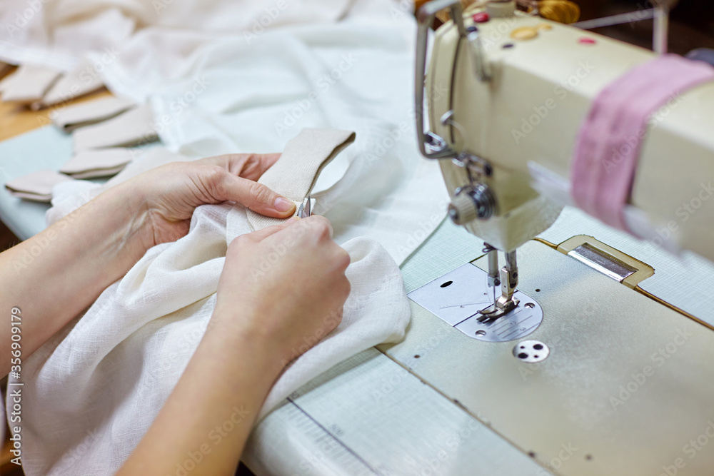 Close up view of cropped female hands sewing white fabric on professional manufacturing machine at seamstress workplace. Tailor hands holding textile sewing item. Soft focus