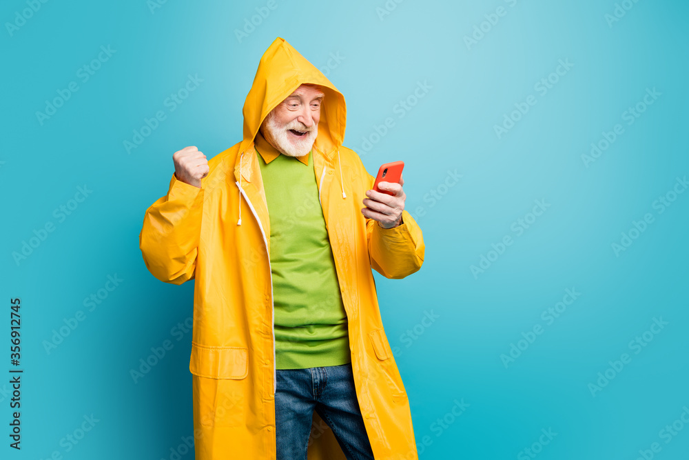Portrait of his he nice cheerful cheery satisfied grey-haired man wearing yellow topcoat using cell fast speed internet connection isolated on bright vivid shine vibrant blue color background