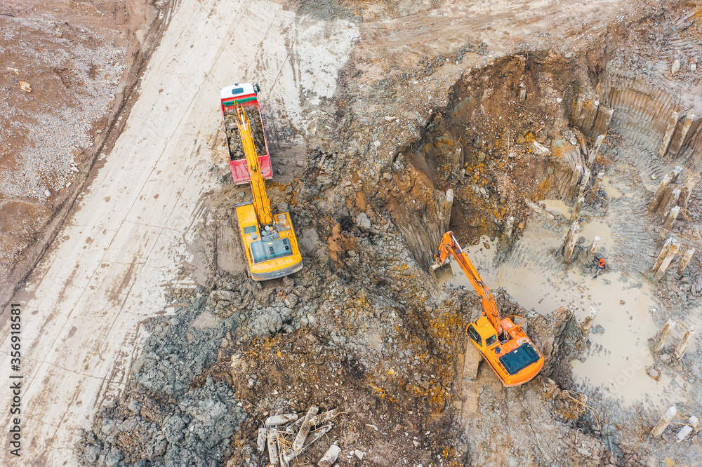 Excavator loading soil into trucks in a foundation pit with piles, aerial top view